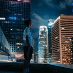 A contemplative young man looks out at Hong Kong's illuminated skyscrapers under a night sky.