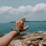 A hand with a watch reaching towards the ocean against a rocky beach under a clear sky.
