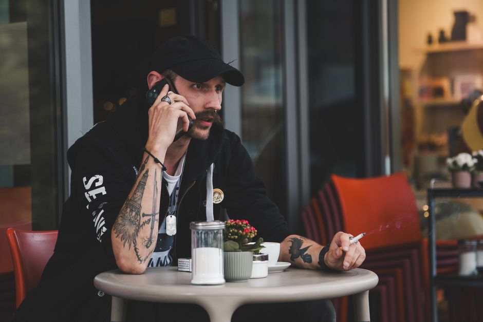 Casual man with tattoos seated at outdoor cafe, talking on phone while smoking.